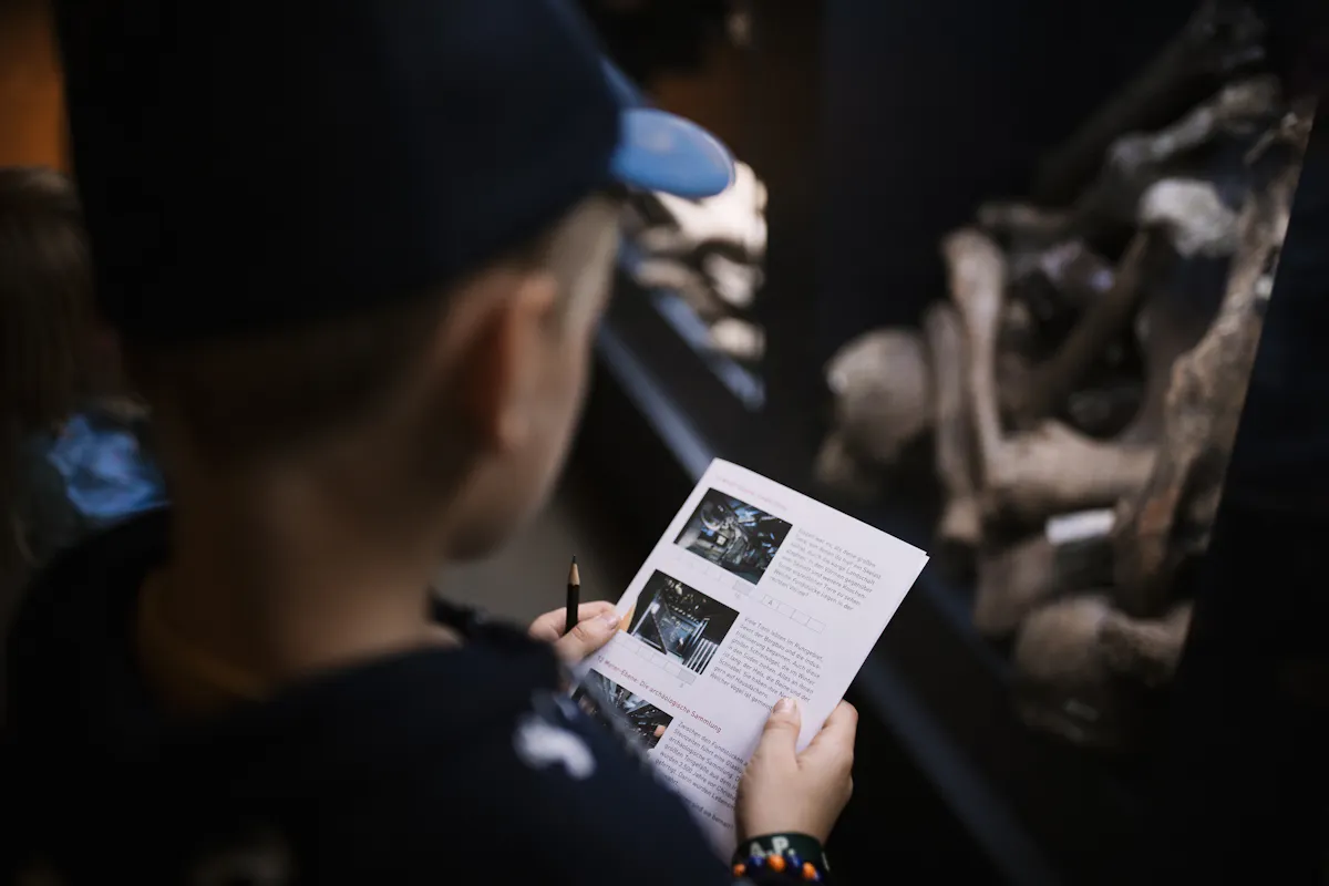 Ein Kind mit dunkler Kappe hält ein Blatt Papier mit Bildern und Texten in der Hand und betrachtet eine Museumsvitrine. In der Vitrine liegen große Fossilien oder Knochenstücke. Das Kind hält einen Stift und scheint an einer Museumsaufgabe oder Rallye teilzunehmen.