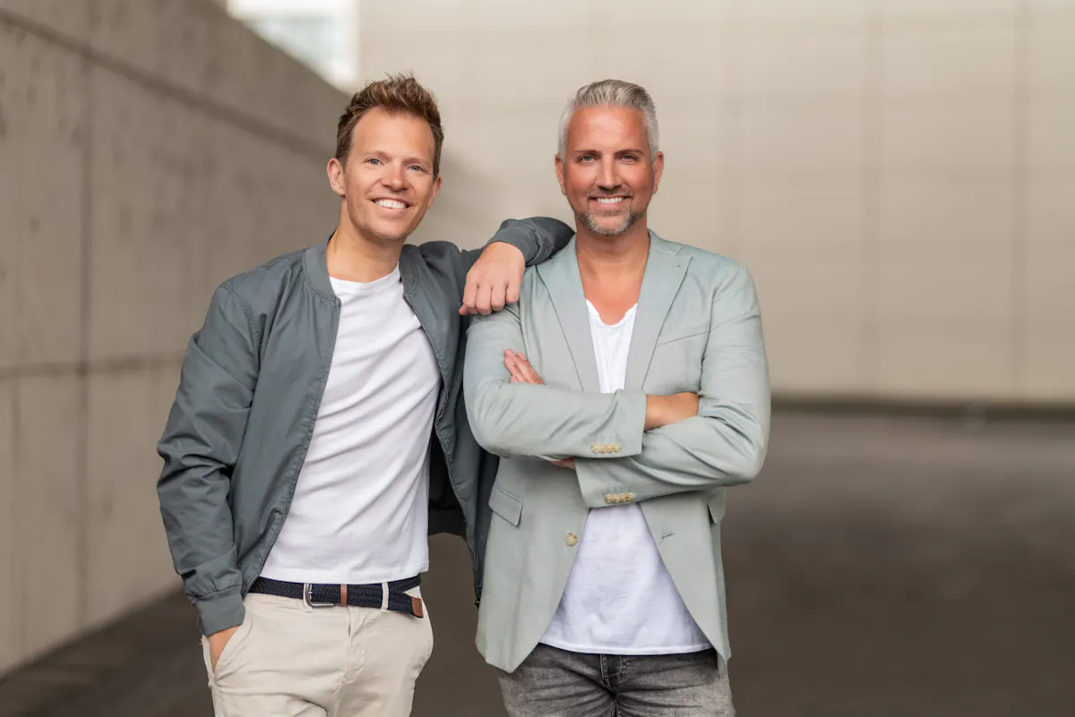 Zwei Männer stehen nebeneinander und lächeln in die Kamera. Der Mann links trägt eine graue Jacke, ein weißes T-Shirt und helle Hosen, mit einer Hand in der Hosentasche und der anderen locker auf der Schulter des anderen Mannes. Der Mann rechts hat graue Haare, trägt einen hellen Blazer über einem weißen T-Shirt und verschränkt die Arme. Der Hintergrund ist eine moderne Betonwand.