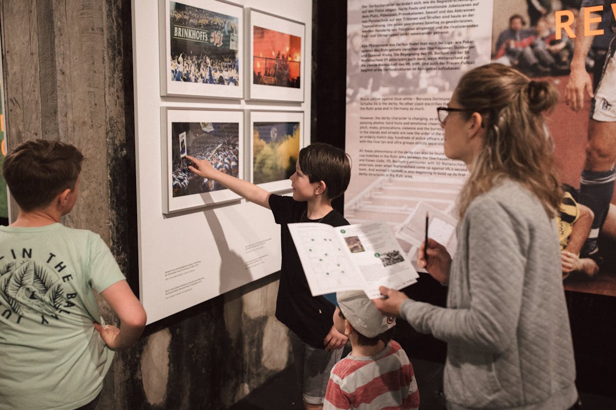 Eine Frau und drei Kinder stehen in einer Ausstellung und betrachten gerahmte Fotos an der Wand, die Fußballfans und Stadionszenen zeigen. Ein Junge zeigt auf eines der Bilder, während die anderen aufmerksam zuhören. Zwei von ihnen halten Blätter mit Aufgaben oder Informationen in den Händen.