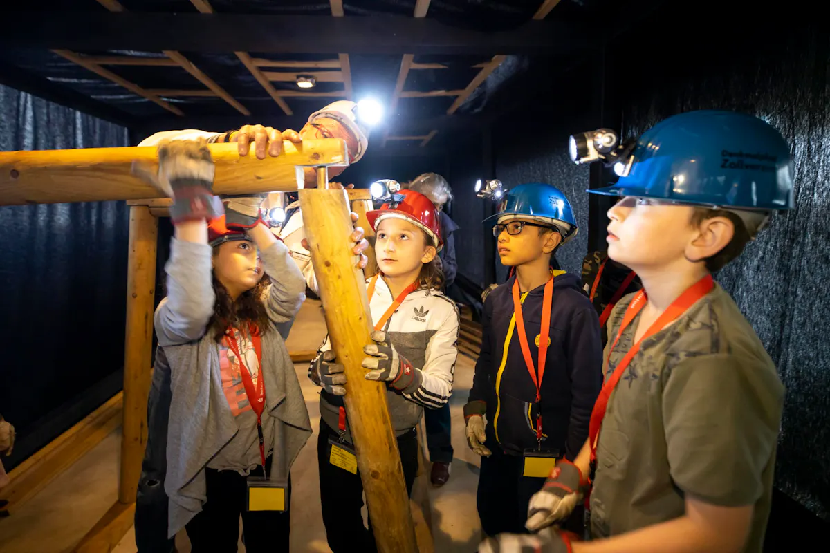 Mehrere Kinder tragen Schutzhelme mit Stirnlampen und bauen gemeinsam mit einem Erwachsenen aus Holzstämmen eine Stützkonstruktion. Sie stehen in einem dunklen Raum, der an einen Bergwerksstollen erinnert. Die Kinder wirken konzentriert und interessiert.
