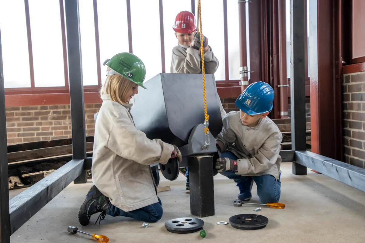 Drei Kinder tragen Schutzhelme und helle Arbeitsjacken. Sie montieren gemeinsam einen kleinen Förderwagen, der mit einem gelben Seil leicht hochgezogen ist. Auf dem Boden liegen Werkzeuge und Schrauben. Die Szene spielt in einer Industriehalle mit Backsteinwänden und Metallträgern.