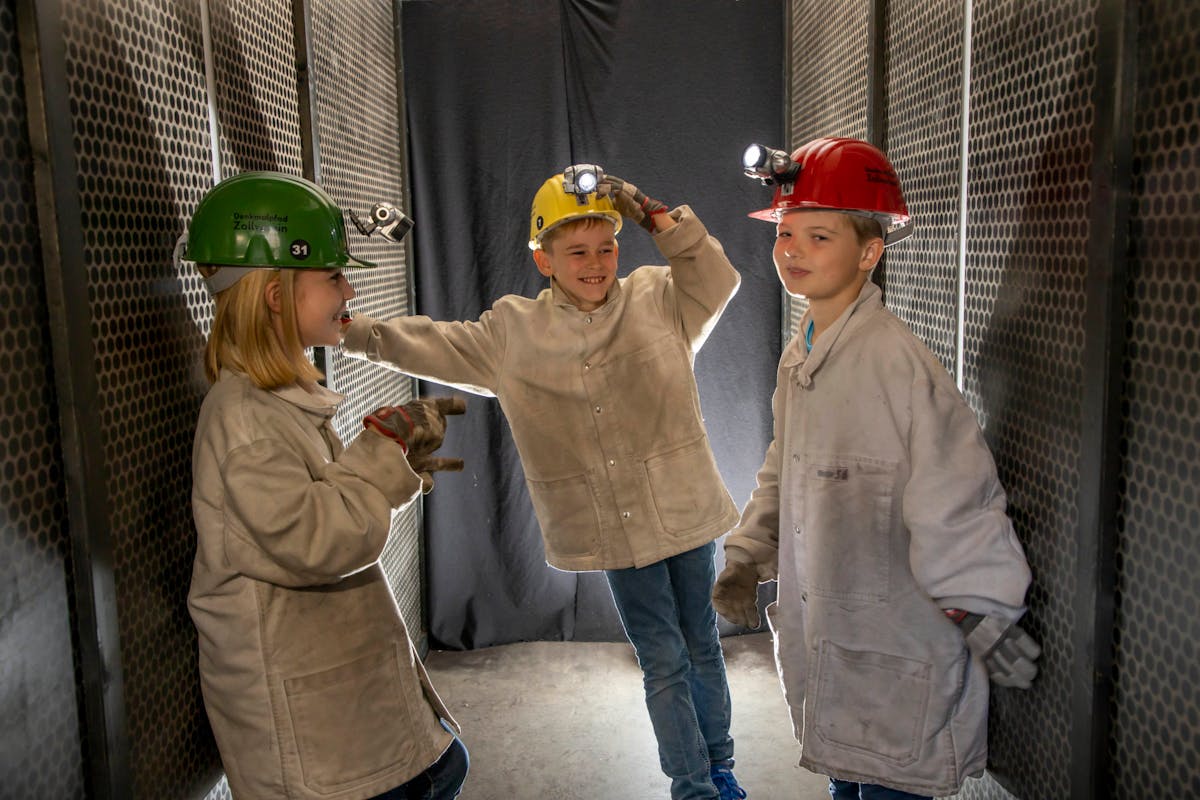 Drei Kinder in Arbeitsjacken und Schutzhelmen mit Stirnlampen stehen lachend in einem schmalen, metallverkleideten Gang. Sie scheinen sich zu amüsieren und spielen ausgelassen, als wären sie junge Bergleute bei einem Erlebnisprogramm.