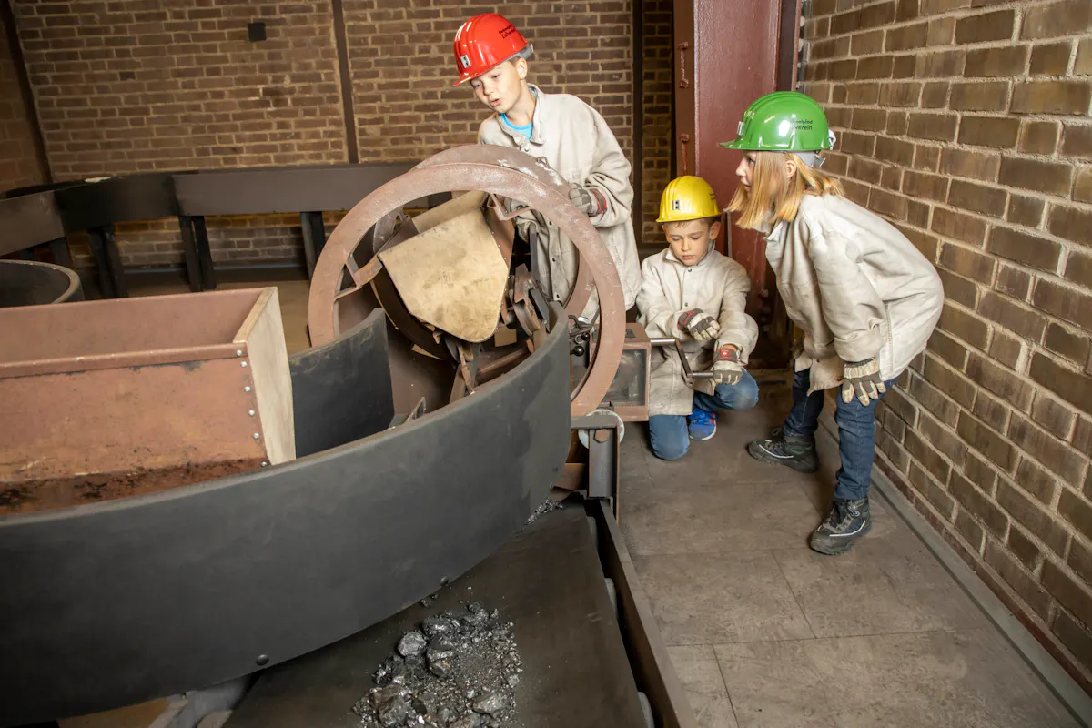 Drei Kinder tragen bunte Schutzhelme und helle Arbeitsjacken. Sie stehen an einer nachgebildeten Industrieanlage mit Förderwagen und untersuchen neugierig die Mechanik. Auf dem Band liegen Stücke dunkler Kohle. Die Szene spielt in einem Backsteinraum.