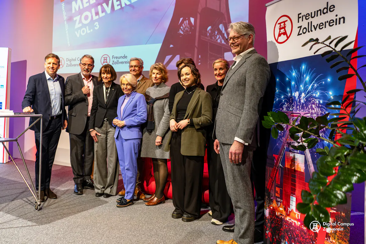 Eine Gruppe von zehn Personen steht lächelnd auf einer Bühne und posiert für ein gemeinsames Foto. Im Hintergrund sind eine Leinwand mit dem Schriftzug Zollverein meets Zollverein Vol. 3 sowie Banner der Freunde Zollverein und des Digital Campus Zollverein zu sehen. Die Stimmung wirkt festlich und professionell.