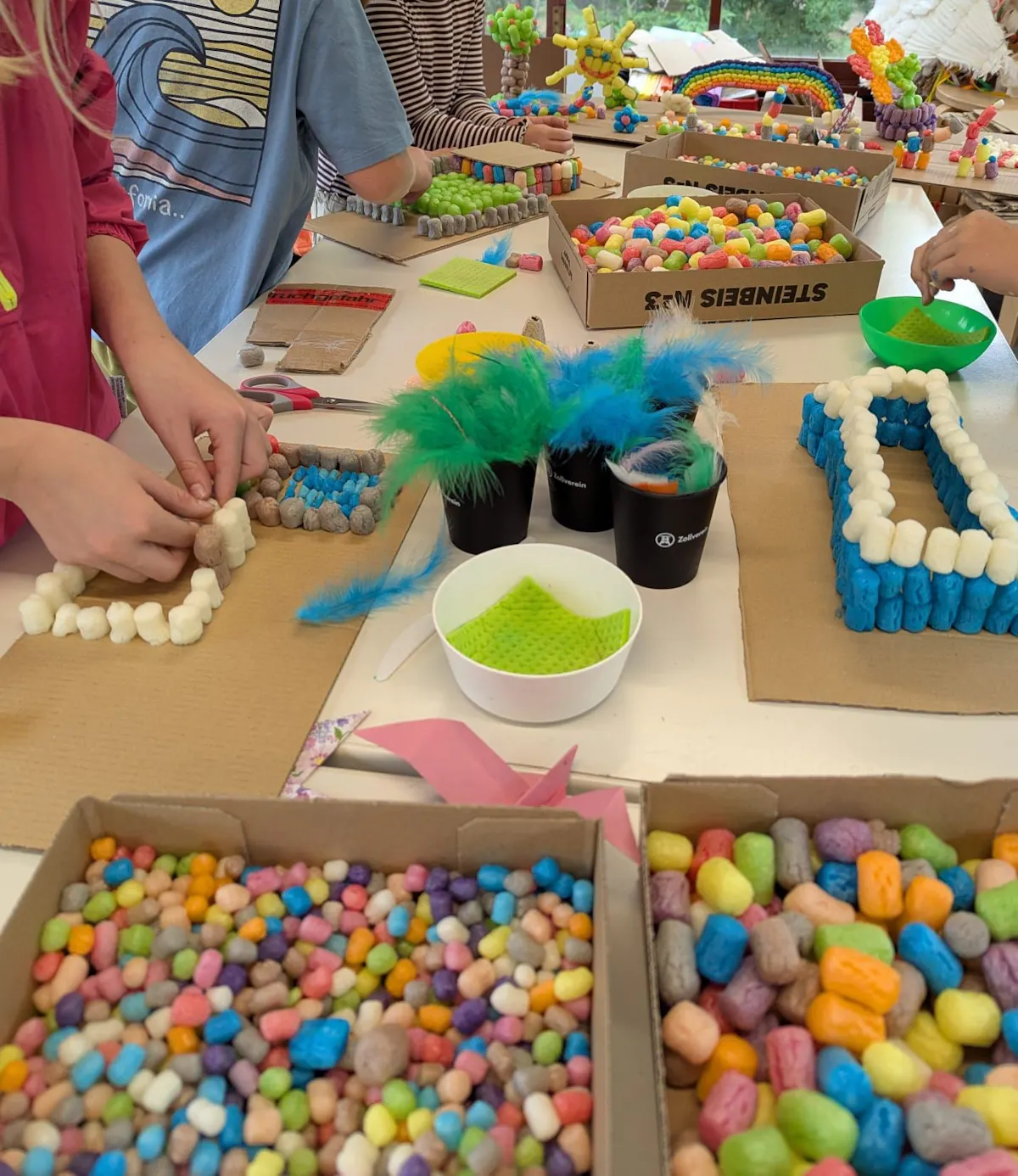 Kinder basteln an einem Tisch mit bunten Materialien. Auf Kartons kleben sie farbige Maisbausteine zu kleinen Bauwerken, daneben liegen Scheren, Schwämme und Federn in kräftigen Farben. Der Tisch ist voller kreativer Bastelprojekte in verschiedenen Stadien.
