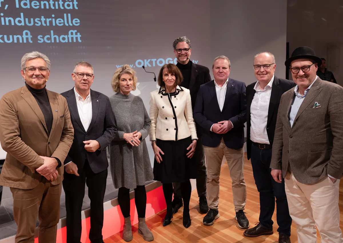 Acht Personen stehen lächelnd für ein Gruppenfoto auf einer Bühne. Sie tragen überwiegend geschäftliche Kleidung. Im Hintergrund sind Teile einer Projektion mit dem Veranstaltungsthema zu Industriekultur, Identität und Innovation sowie der Schriftzug OKTOGON.tv zu sehen.