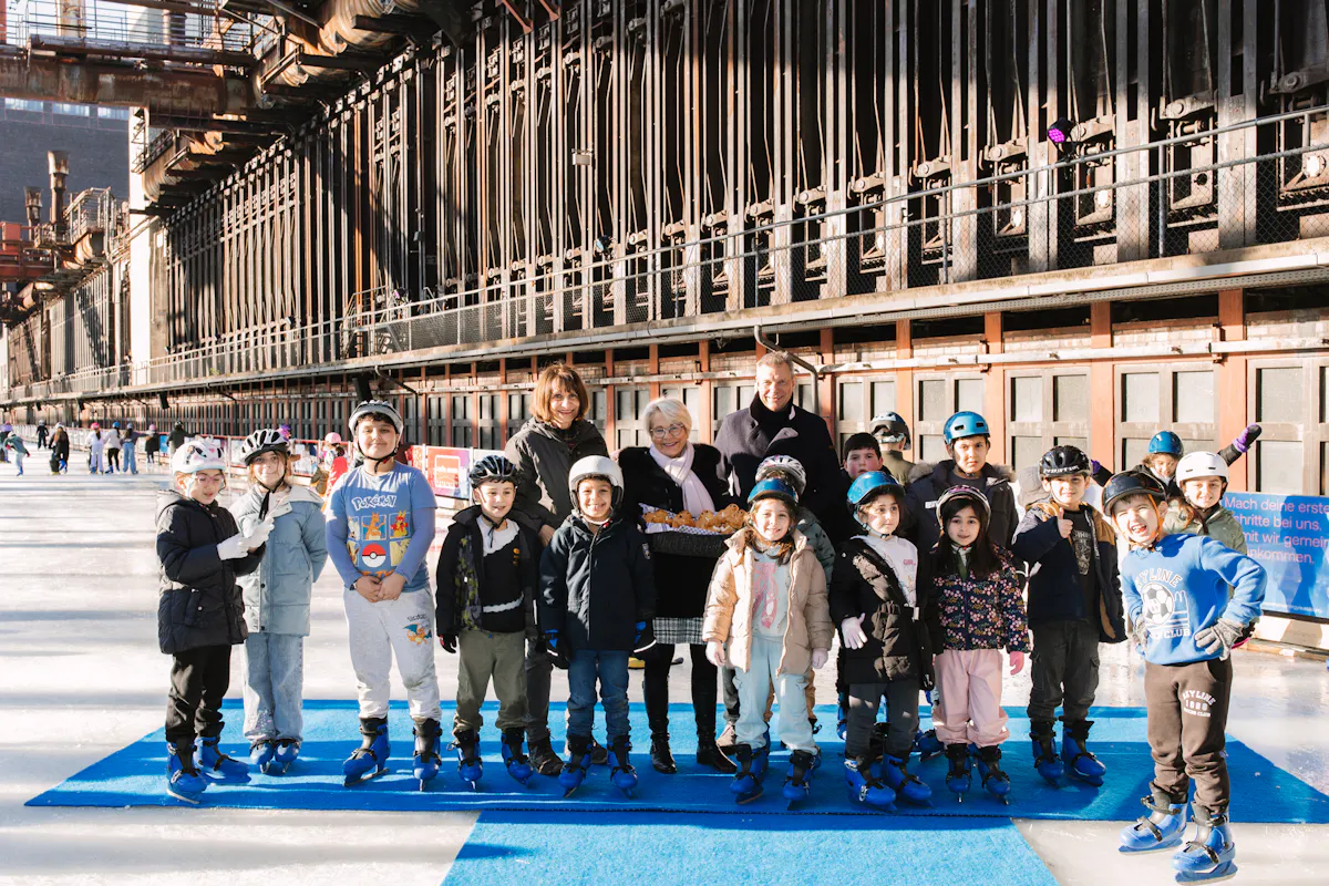 Eine Gruppe von Kindern mit Schlittschuhen und Helmen steht gemeinsam mit mehreren Erwachsenen auf einer Eisbahn im Freien. Im Hintergrund verläuft die lange industrielle Fassade der Zollverein-Anlage. Die Szene wirkt wie ein Gruppenfoto bei einem Eislaufangebot für Kinder.