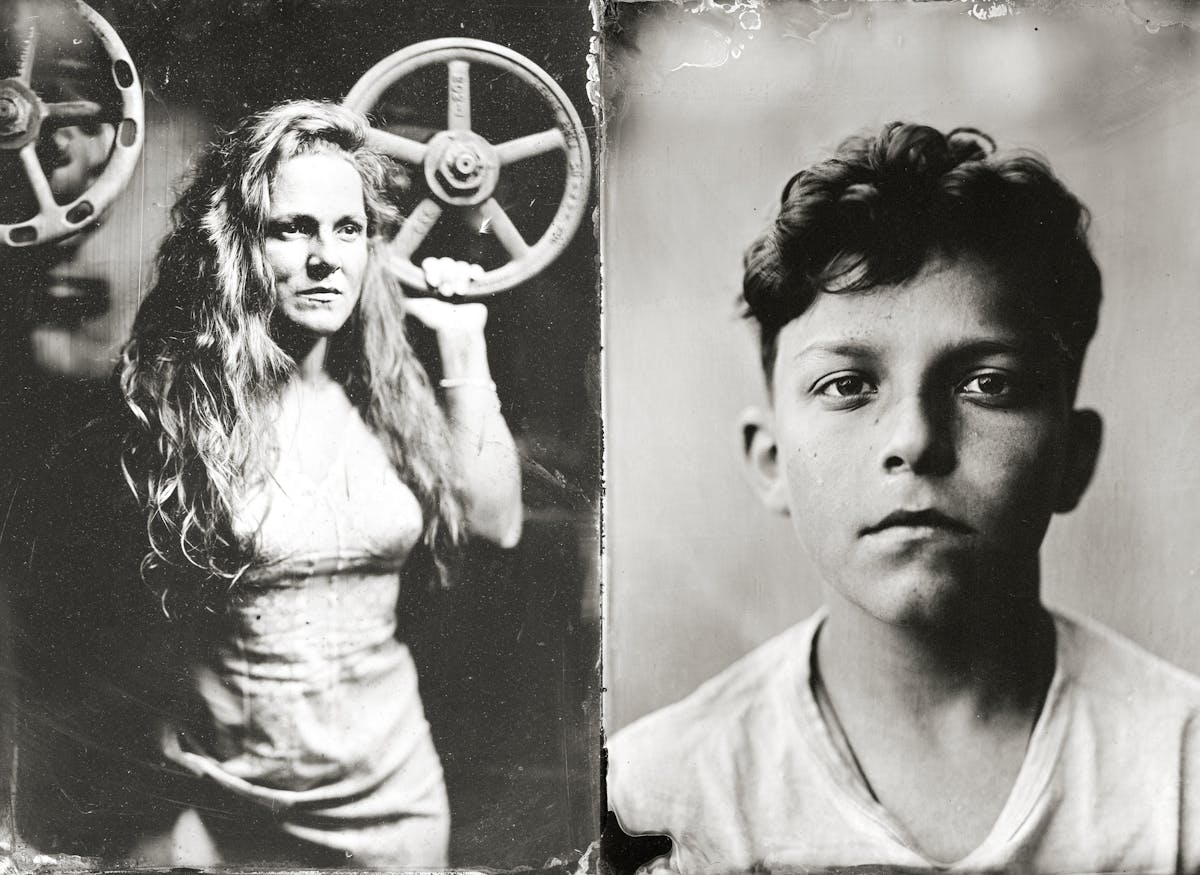 Zwei nebeneinander angeordnete Porträts im Stil einer historischen Schwarz-weiß-Fotografie. Links steht eine Person mit langen, gewellten Haaren vor einem großen, runden Metallrad, das an eine Maschine erinnert. Die Person trägt ein ärmelloses, eng anliegendes Kleid. Rechts ist eine Person mit kurzem, gewelltem Haar und einem weißen T-Shirt zu sehen. Der Hintergrund beider Bilder ist unscharf und erzeugt einen nostalgischen Effekt.