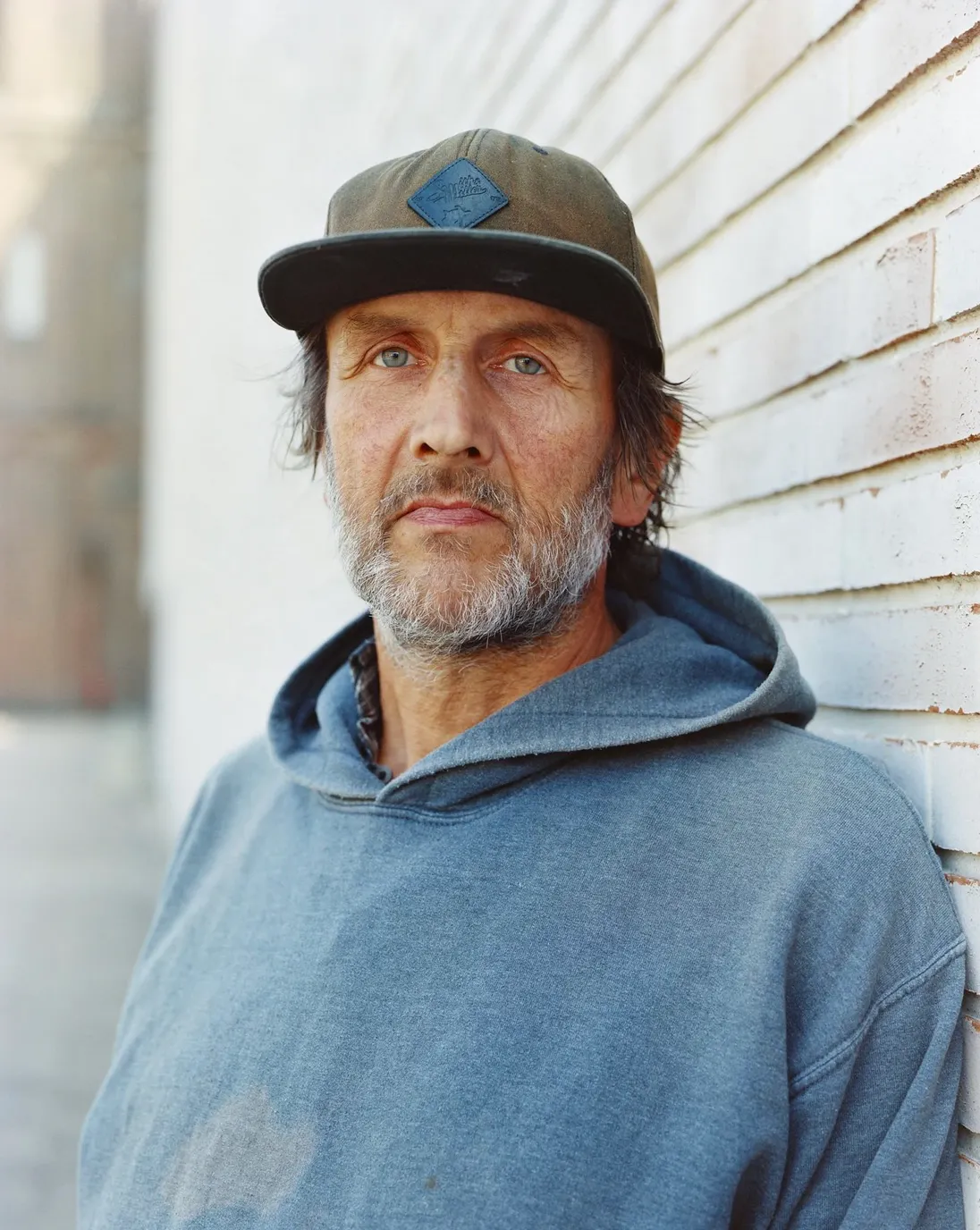 Eine Person trägt einen grauen Kapuzenpullover und eine olivgrüne Baseballkappe mit einem abgesetzten Emblem auf der Vorderseite. Die Person steht vor einer hellen, gemauerten Wand, die im rechten Bildbereich verläuft. Der Hintergrund ist unscharf und zeigt eine städtische Umgebung.