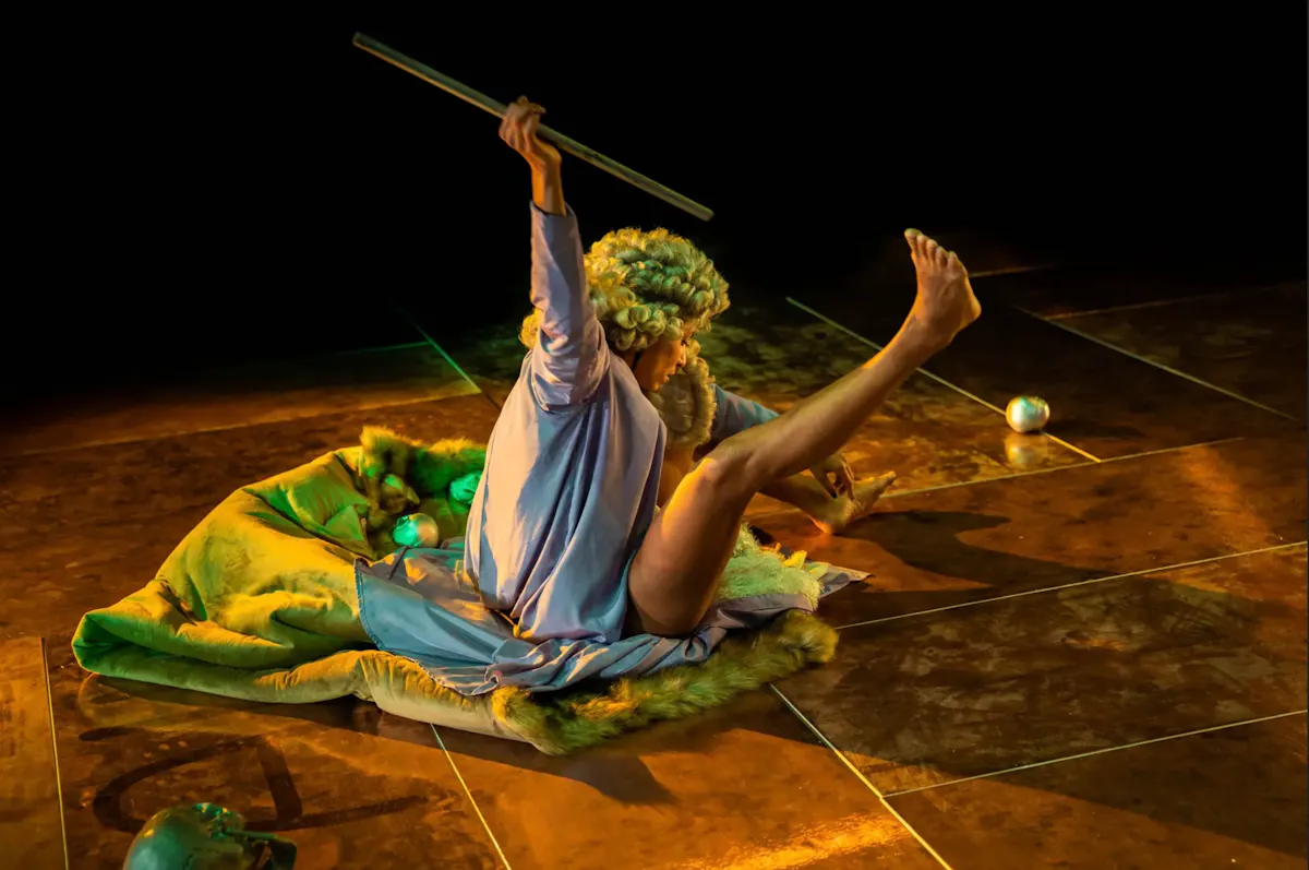 Eine Person mit lockiger, heller Perücke sitzt auf einem mit Fell bedeckten Untergrund. Sie trägt ein helles, locker fallendes Gewand und hält einen langen Stab in einer erhobenen Hand. Ein Bein ist angewinkelt und in die Höhe gestreckt. Die Szene ist in warmes, gelbliches Licht getaucht. Auf dem Boden liegen mehrere glänzende, eiförmige Objekte verteilt.