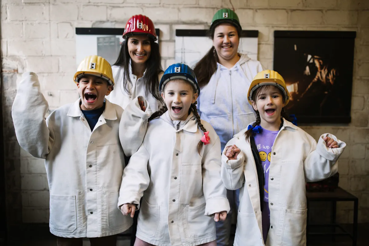 Drei Kinder und zwei Erwachsene stehen in einem Ausstellungsraum und schauen fröhlich in die Kamera. Die Kinder tragen weiße Arbeitsjacken und gelbe beziehungsweise blaue Schutzhelme, die Erwachsenen im Hintergrund rote und grüne Schutzhelme. Alle wirken ausgelassen, mehrere haben die Münder geöffnet oder die Fäuste erhoben. Hinter ihnen sind helle Steinwände und dunkle Ausstellungstafeln zu sehen.