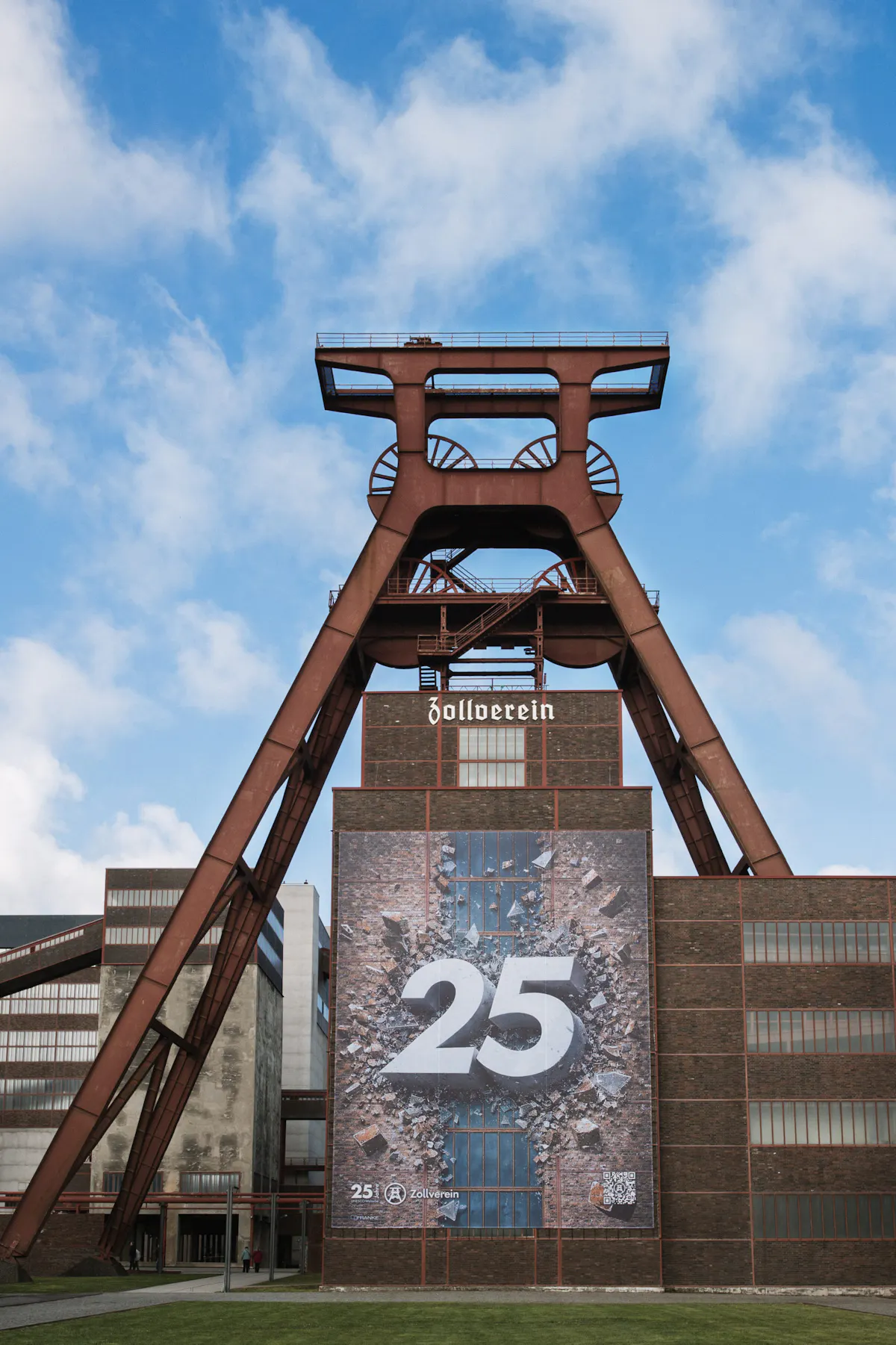 Frontalansicht des Fördergerüsts der Zeche Zollverein vor einem Backsteingebäude unter blauem Himmel mit lockeren Wolken. Auf der Gebäudefassade hängt ein großes Banner mit der Zahl 25 in Weiß vor einer aufbrechenden, steinartigen Struktur im Stil der Zollverein-Fassaden. Im Vordergrund liegt eine Rasenfläche, seitlich rahmen weitere Industriegebäude die Szene ein.
