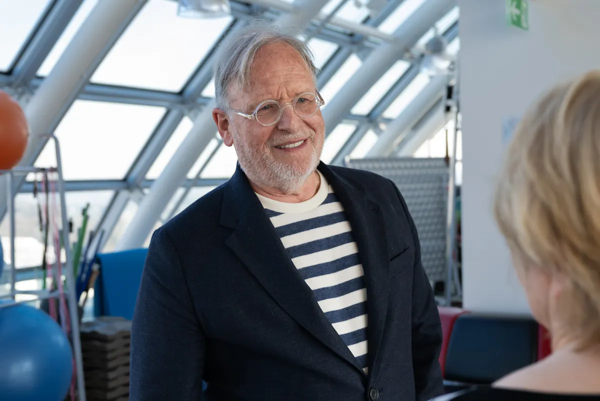 Eine Person trägt ein dunkelblaues Sakko über einem weiß-blau gestreiften Pullover. Im Hintergrund sind große Fenster mit Metallstreben, bunte Gymnastikbälle und gestapelte Matten zu sehen. Eine weitere Person mit blondem, kurzem Haar steht im Vordergrund mit dem Rücken zur Kamera.