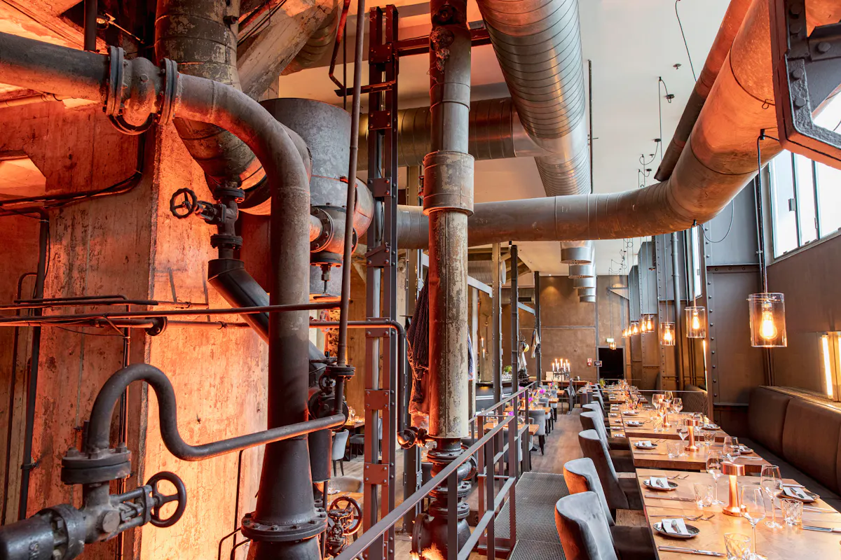 Restaurant mit industriellem Ambiente in einer ehemaligen Industriehalle. Links verlaufen große Rohre, Ventile und Metallstützen entlang der Wand, warm orange beleuchtet. Rechts stehen lange gedeckte Tische mit Gläsern, Tellern, Besteck und kleinen Lampen. Hohe Fenster, sichtbare Lüftungsrohre und Hängelampen prägen den Raum.