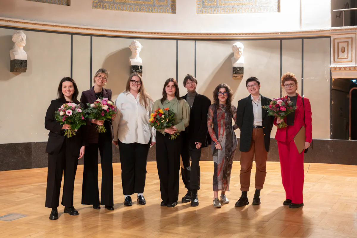 Acht Personen stehen nebeneinander auf einer Bühne oder in einem Festsaal und schauen in die Kamera. Mehrere von ihnen halten bunte Blumensträuße, die Person rechts trägt einen auffälligen roten Anzug und hält zusätzlich eine Mappe. Im Hintergrund sind helle Wände, Büsten und dekorative Verzierungen zu sehen. Der Raum wirkt feierlich und wird warm beleuchtet.