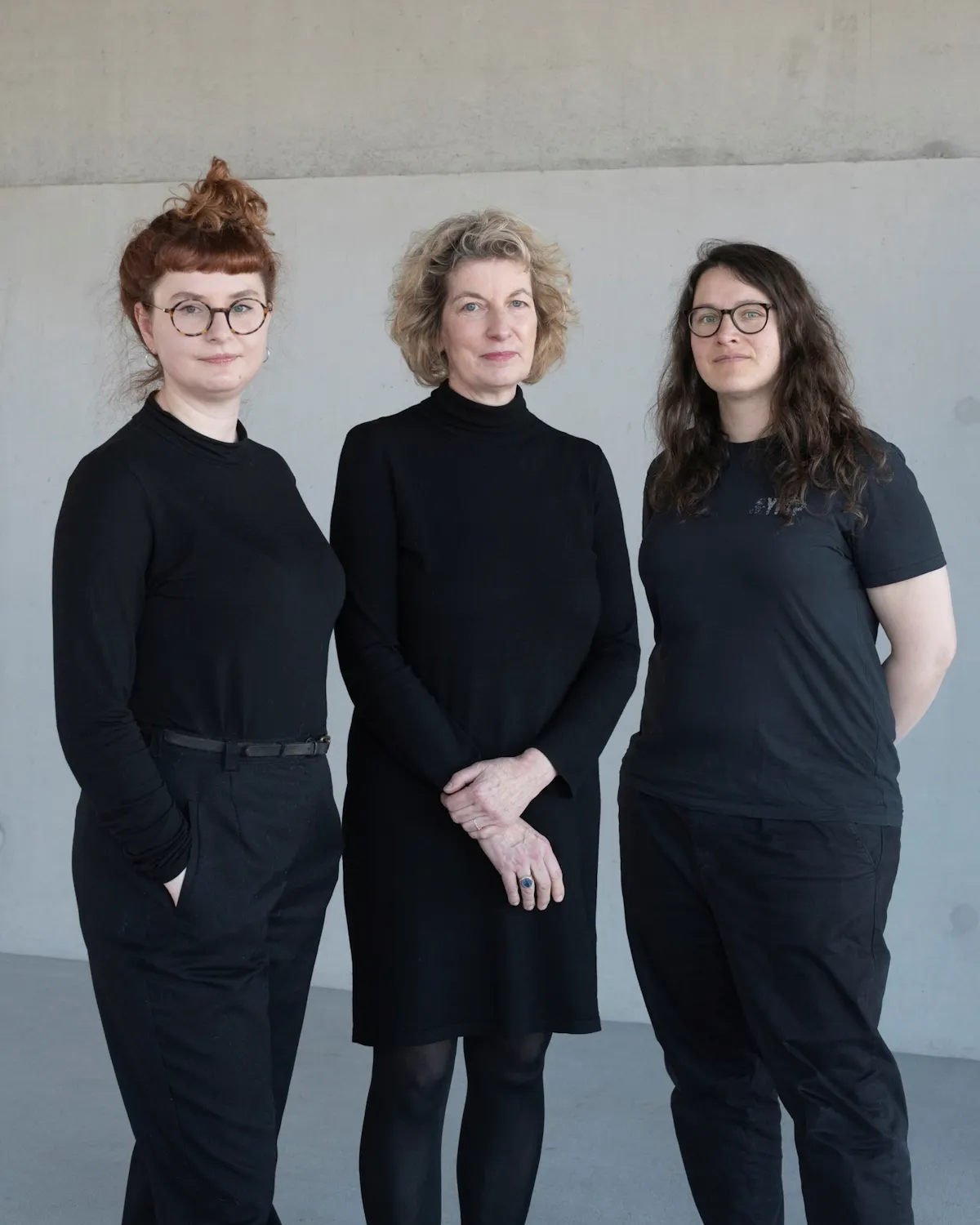 Drei Personen stehen nebeneinander vor einer hellgrauen Wand. Alle tragen schwarze Kleidung. Die mittlere Person hält die Hände vor dem Körper, während die beiden anderen die Arme seitlich locker hängen lassen. Die Gesichter sind unkenntlich gemacht.