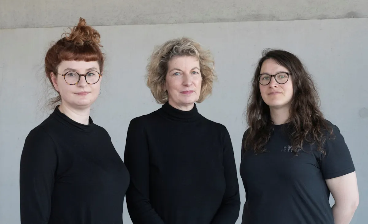 Drei Personen stehen nebeneinander vor einer hellgrauen Wand. Alle tragen schwarze Kleidung. Die mittlere Person hält die Hände vor dem Körper, während die beiden anderen die Arme seitlich locker hängen lassen. Die Gesichter sind unkenntlich gemacht.