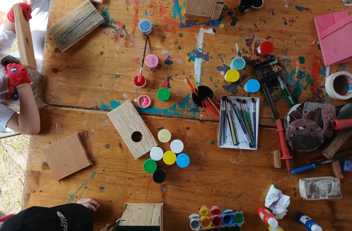 Auf einer hölzernen Tischplatte liegen zahlreiche bunte Farbdosen mit geöffneten Deckeln, mehrere Pinsel, Holzstücke mit Bohrlöchern, ein Schraubstock, Schleifpapier und weitere Bastelmaterialien. Zwei Kinderarme sind am unteren und linken Bildrand zu sehen, eines der Kinder trägt einen roten Handschuh. Die Tischoberfläche ist mit Farbflecken übersät.