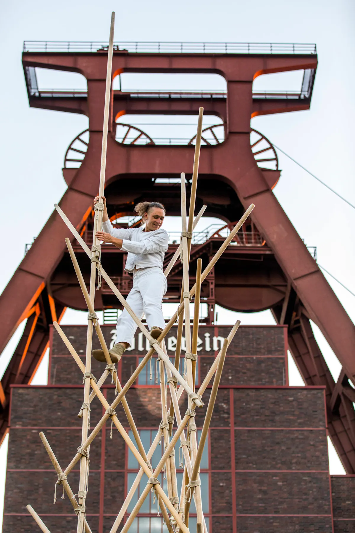 Eine Person in weißer Kleidung steht auf einer hohen, aus Bambusstäben gebauten Konstruktion. Im Hintergrund ist das markante Fördergerüst der Zeche Zollverein aus rostbraunem Stahl zu sehen. Das Gebäude darunter besteht aus dunklen Ziegelsteinen, auf denen der Schriftzug Zollverein angebracht ist. Der Himmel ist hell und wolkenlos.