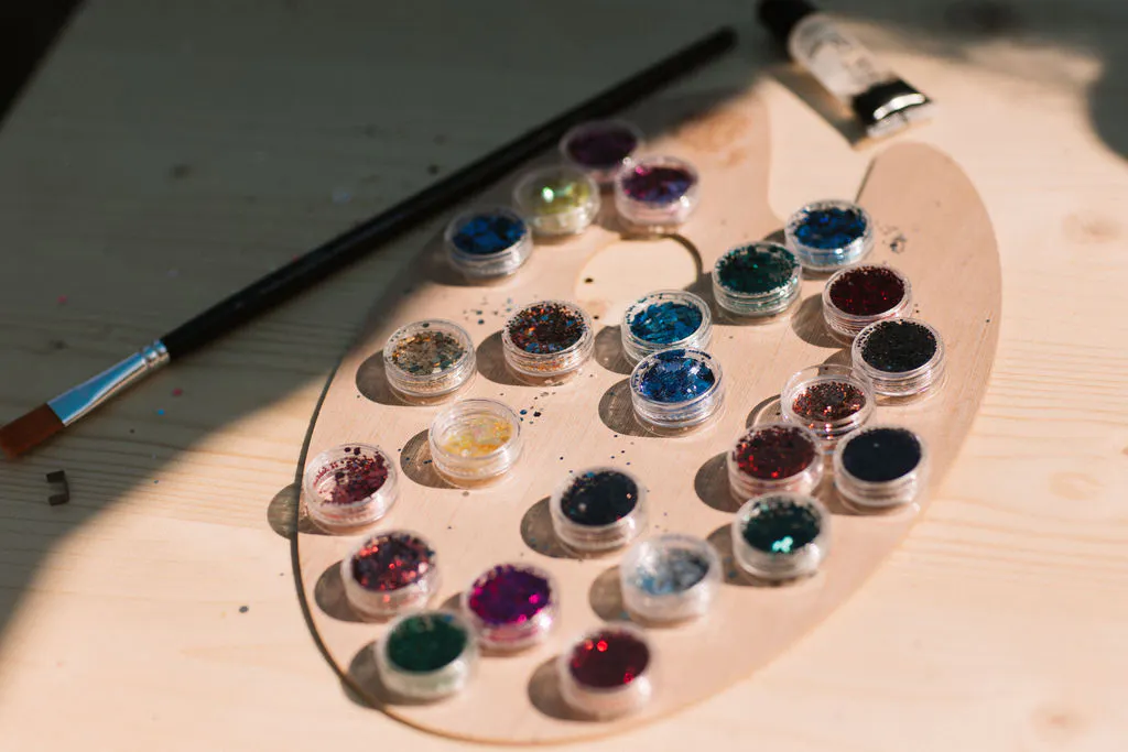 Eine ovale Holzpalette liegt auf einer hellen Tischplatte. Darauf stehen zahlreiche kleine, runde Döschen mit glitzerndem Farbpulver in verschiedenen Farben. Ein Pinsel und eine Tube mit Farbresten liegen daneben. Das Licht fällt schräg auf die Szene und wirft deutliche Schatten.