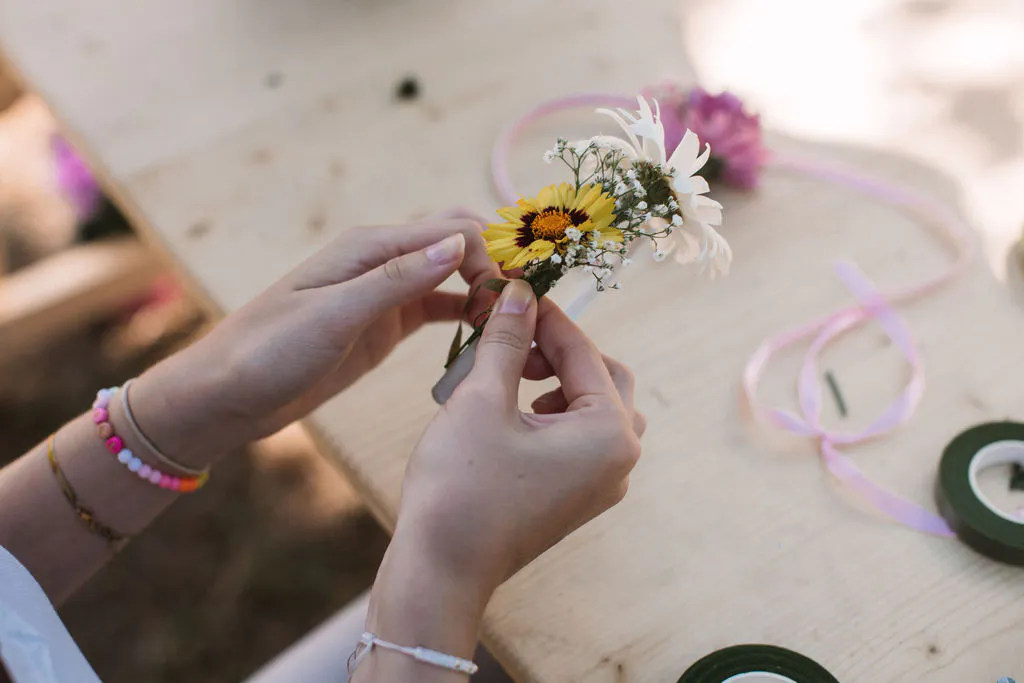 Zwei Hände halten einen kleinen Strauß aus gelben, weißen und violetten Blüten sowie etwas Schleierkraut. Auf dem hellen Holztisch liegen rosafarbenes Band, eine Rolle grünes Floristenband und weitere Blumen. Die Person trägt bunte Perlenarmbänder und ein weißes Oberteil.