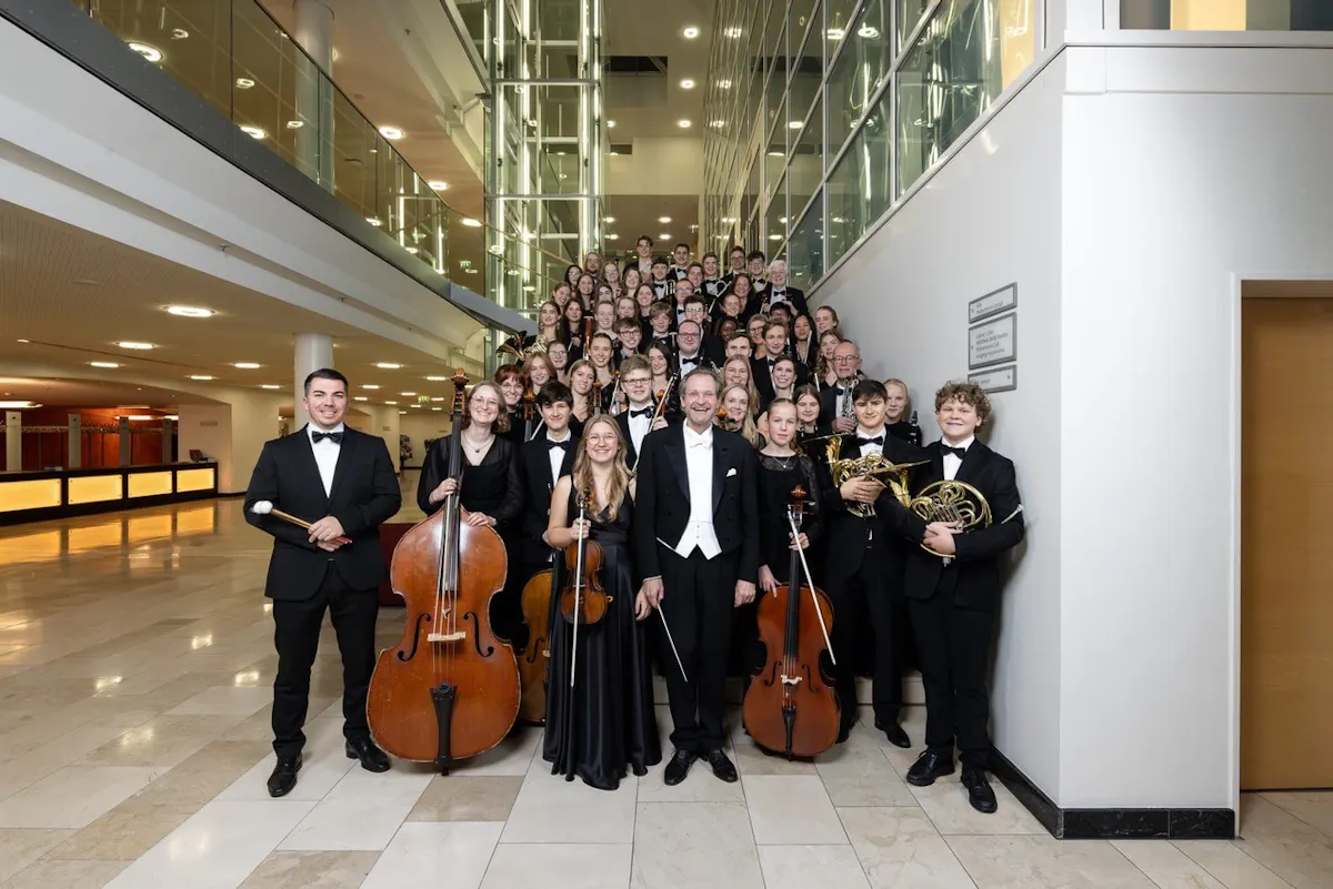 Gruppenfoto eines großen Orchesters oder Ensembles in einem modernen Foyer. Viele Musikerinnen und Musiker in festlicher schwarzer Kleidung stehen dicht gedrängt auf einer Treppe und davor. Im Vordergrund halten mehrere Personen Instrumente wie Cello, Violine, Waldhorn und Schlagzeugschlägel. In der Mitte steht ein Dirigent im Frack und lächelt in die Kamera. Die Umgebung ist hell, mit Glasgeländern, Aufzugsschacht und hohen Innenräumen.