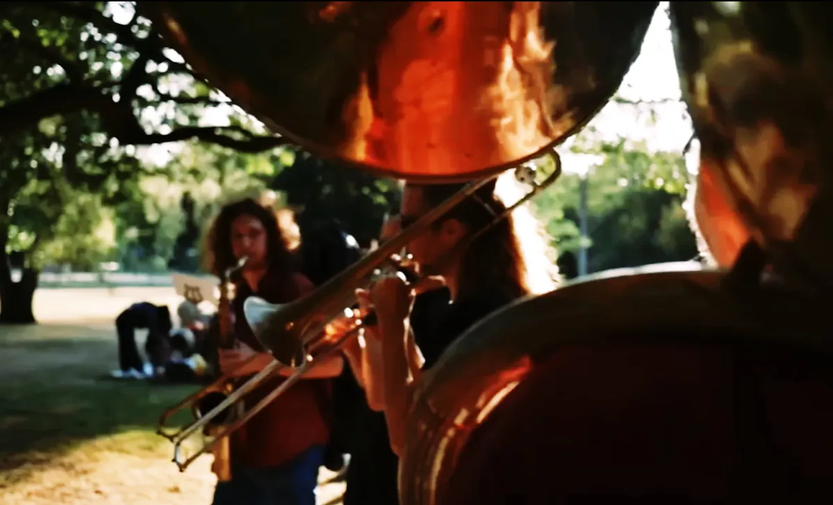 Nahaufnahme einer musizierenden Gruppe im Freien bei warmem Sonnenlicht. Im Vordergrund sind große glänzende Blasinstrumente zu sehen, darunter eine Posaune und ein großes Blechblasinstrument. Dahinter spielen weitere Musikerinnen und Musiker im unscharfen Hintergrund unter Bäumen. Die Szene wirkt sommerlich, lebendig und atmosphärisch.