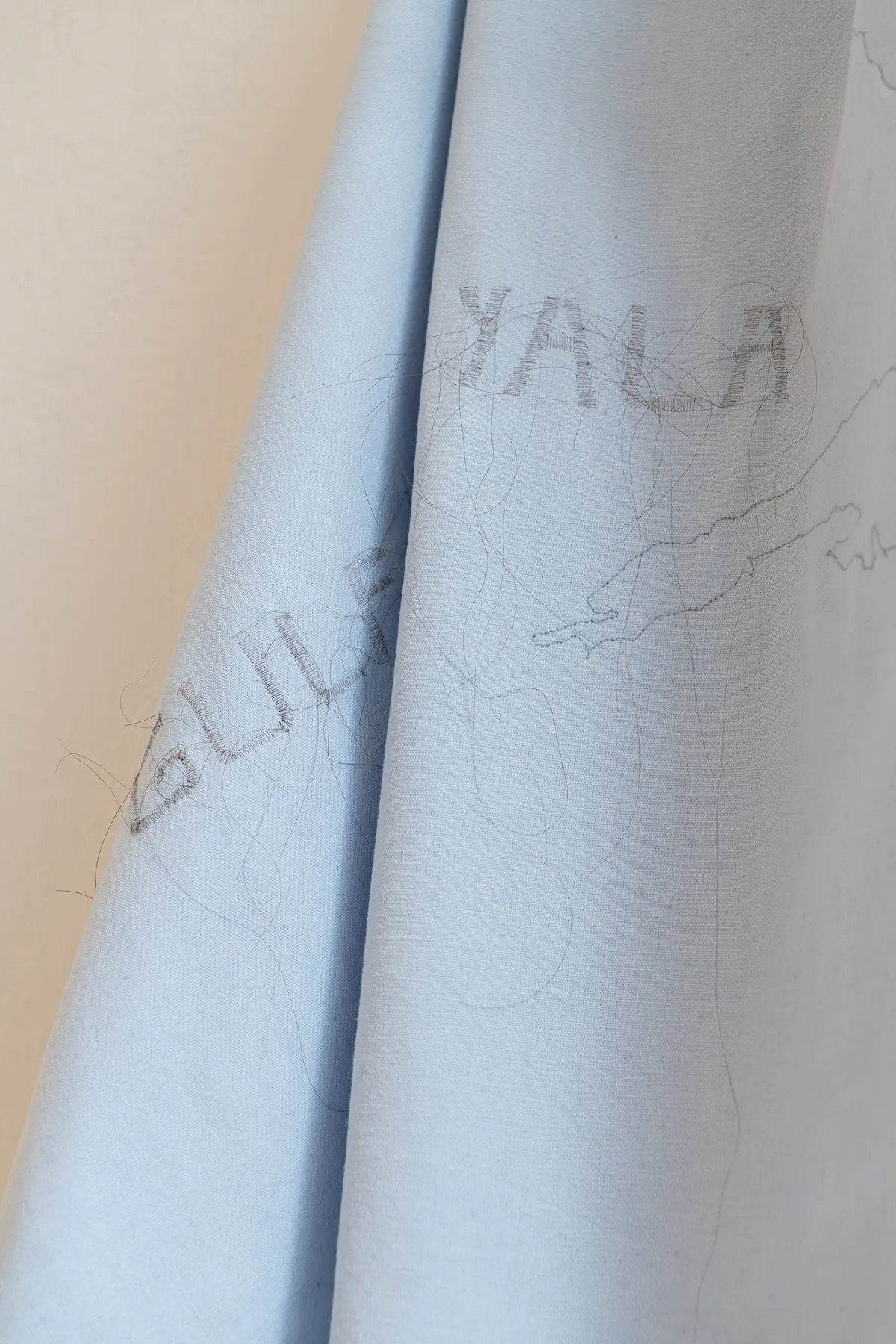 Ein hellblauer, leicht glänzender Stoff hängt in weichen Falten. Auf der Oberfläche sind feine, dunkle Linien und Umrisse zu erkennen, die an eine Landkarte erinnern. Teilweise ist der Schriftzug 'GULF YALA' in klaren Großbuchstaben sichtbar. Die Linien wirken wie mit Faden oder dünnem Garn aufgestickt.