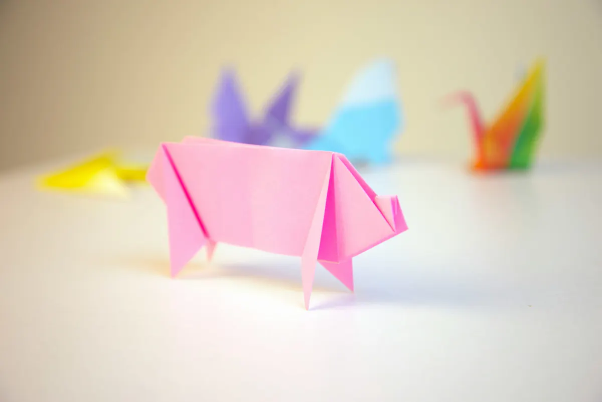 Ein rosa Origami-Schwein steht im Vordergrund auf einer weißen Fläche. Im unscharfen Hintergrund sind weitere Origami-Figuren in verschiedenen Farben zu erkennen.