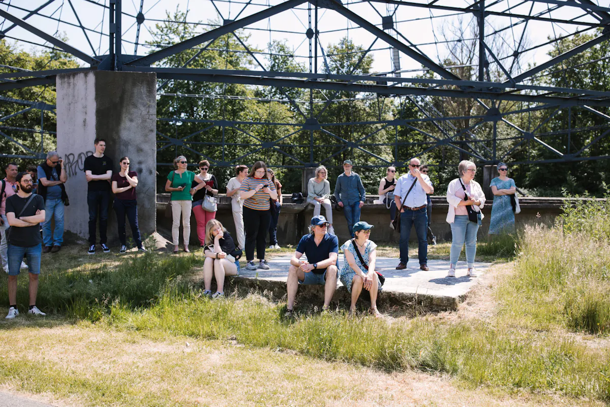 Gruppe von Menschen bei einer Führung im Grünen auf dem Gelände einer ehemaligen Industrieanlage. Einige Personen stehen, andere sitzen auf einer niedrigen Betonfläche. Im Hintergrund ist eine große Stahlkonstruktion zu sehen, dahinter Bäume und Sträucher. Die Szene wirkt sonnig und informell, mit viel Gras und überwachsener Industriekulisse.