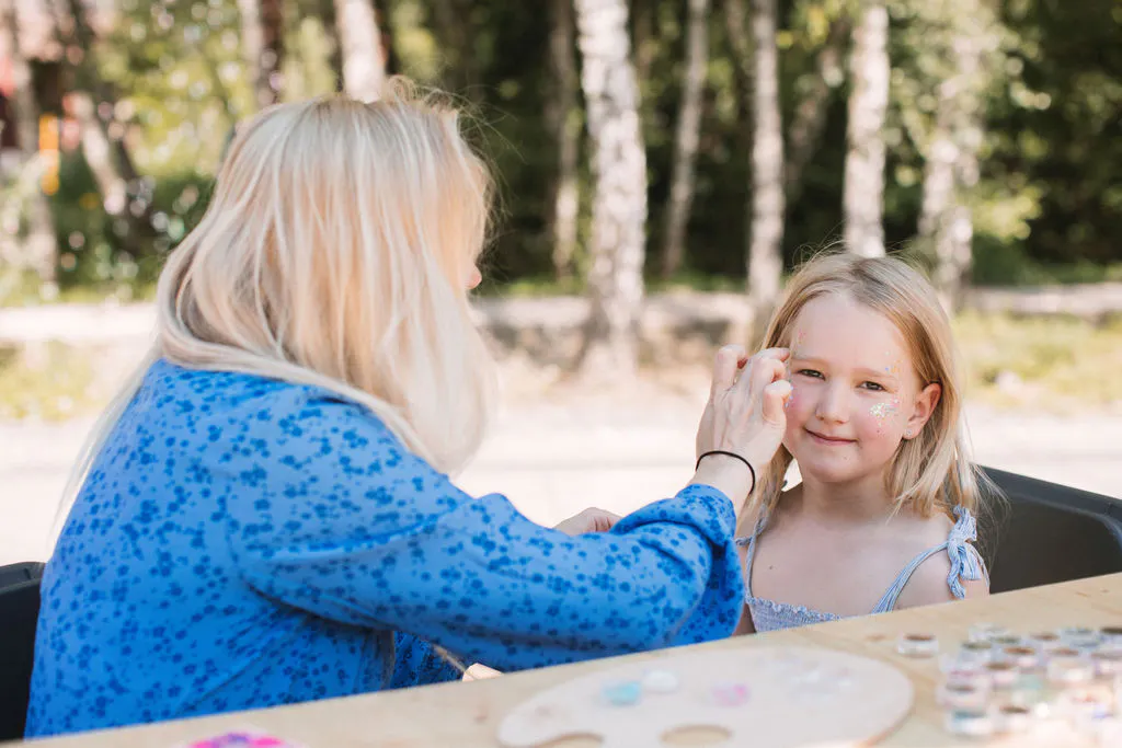 Eine Frau mit langen, blonden Haaren trägt eine hellblaue Bluse mit floralem Muster und bemalt das Gesicht eines Kindes. Das Kind sitzt ihr gegenüber an einem Holztisch im Freien und trägt ein hellblaues Oberteil mit schmalen Trägern. Auf dem Tisch liegen mehrere kleine Dosen mit Farben sowie eine runde Palette. Im Hintergrund sind unscharf Bäume und grüne Vegetation zu erkennen.