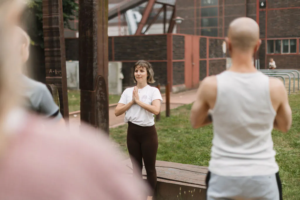 Outdoor-Yogakurs auf dem Gelände der Zeche Zollverein. Eine Kursleiterin steht mit vor der Brust gefalteten Händen auf einer Holzfläche und blickt zu den Teilnehmenden. Im Vordergrund sind zwei Personen unscharf von hinten zu sehen. Im Hintergrund befinden sich Wiesen, Stahlträger und Backsteingebäude der Industrieanlage.
