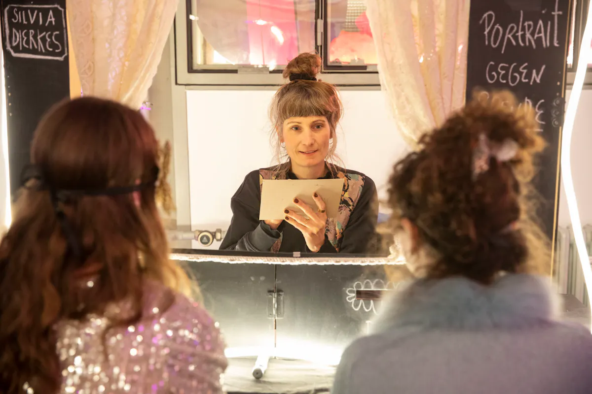 Zwei Personen mit glitzernden Oberteilen und Masken sitzen an einem Tisch, ihnen gegenüber eine Person mit hochgesteckten Haaren, die ein Blatt Papier in den Händen hält. Im Hintergrund sind helle Vorhänge und ein Fenster zu sehen. Links und rechts am Tisch stehen Tafeln mit handgeschriebener weißer Schrift.