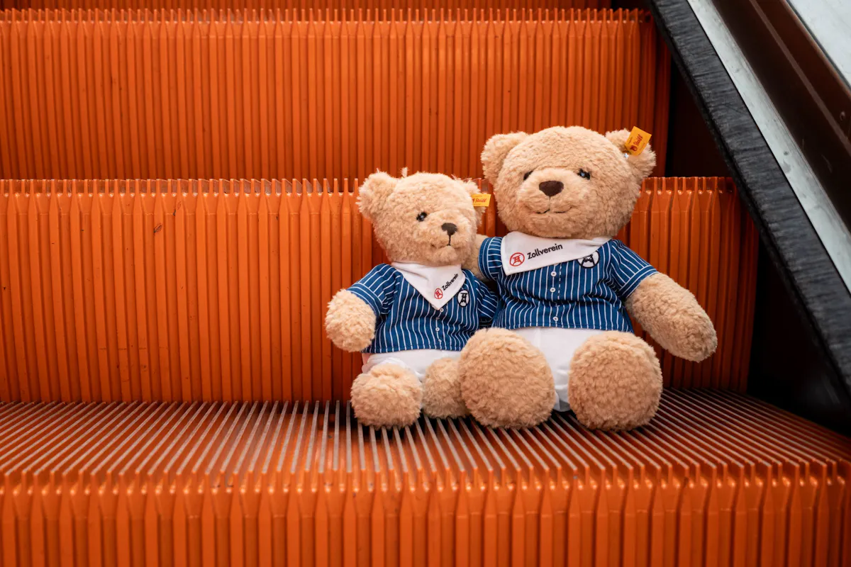 Zwei hellbraune Steiff-Teddybären sitzen nebeneinander auf orangefarbenen Rolltreppenstufen. Beide tragen blau-weiß gestreifte Hemden und weiße Halstücher mit Zollverein-Logo. Der kleinere Bär sitzt links, der größere rechts. Die kräftig orangefarbene Rolltreppe bildet einen auffälligen Hintergrund und betont die beiden Plüschtiere als Motiv