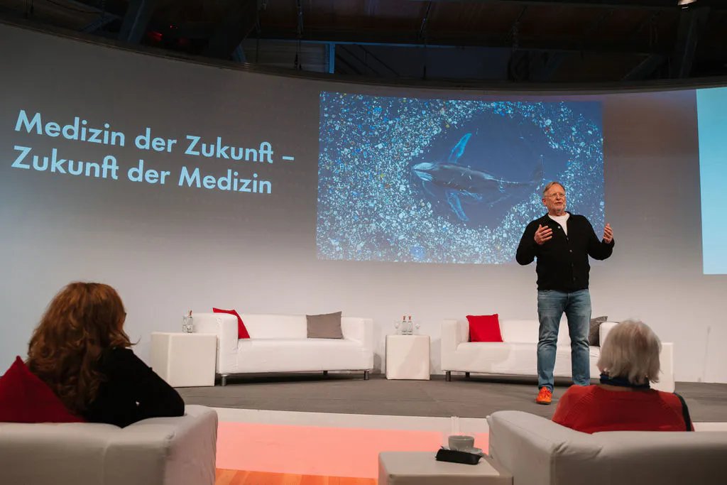 Vortragsszene in einem Veranstaltungsraum mit weißen Sofas vor einer großen Projektionswand. Auf der Bühne steht ein Mann in schwarzer Strickjacke und Jeans und spricht gestikulierend zum Publikum. Auf der Projektion steht Medizin der Zukunft – Zukunft der Medizin, daneben ist ein Bild mit einer Unterwasser- oder Meeresaufnahme zu sehen. Vorne sitzen einzelne Gäste und verfolgen den Vortrag.