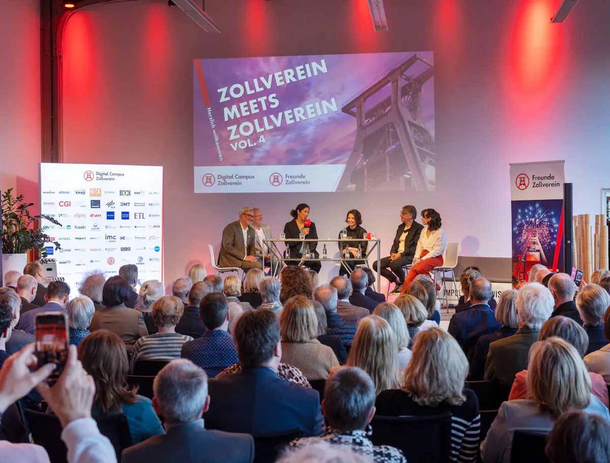Podiumsgespräch im Rahmen von Zollverein meets Zollverein Vol. 4 vor großem Publikum. Auf der Bühne sitzen mehrere Gesprächsteilnehmende an einem hohen Tisch, eine Frau spricht in ein rotes Mikrofon. Dahinter zeigt eine große Projektion den Veranstaltungstitel sowie die Logos von Digital Campus Zollverein und Freunde Zollverein. Links steht eine Fotowand mit Partnerlogos, rechts ein Banner der Freunde Zollverein. Rote Lichtakzente und ein dicht besetzter Saal prägen die festliche Atmosphäre.