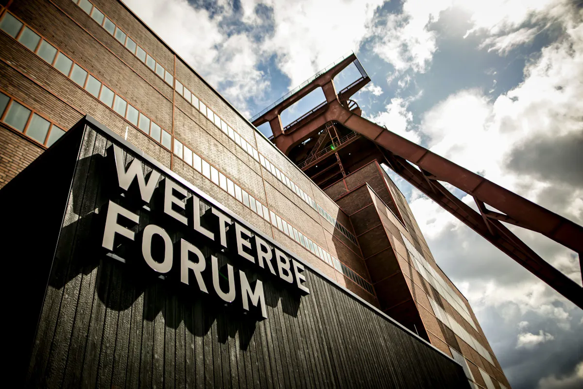 Dramatische Außenaufnahme des Welterbe Forums auf dem Gelände der Zeche Zollverein aus niedriger Perspektive. Im Vordergrund steht der große Schriftzug WELTERBE FORUM an einer dunklen Fassade. Dahinter ragt das rote Doppelbock-Fördergerüst neben einem hohen Backsteingebäude in den wolkigen Himmel.