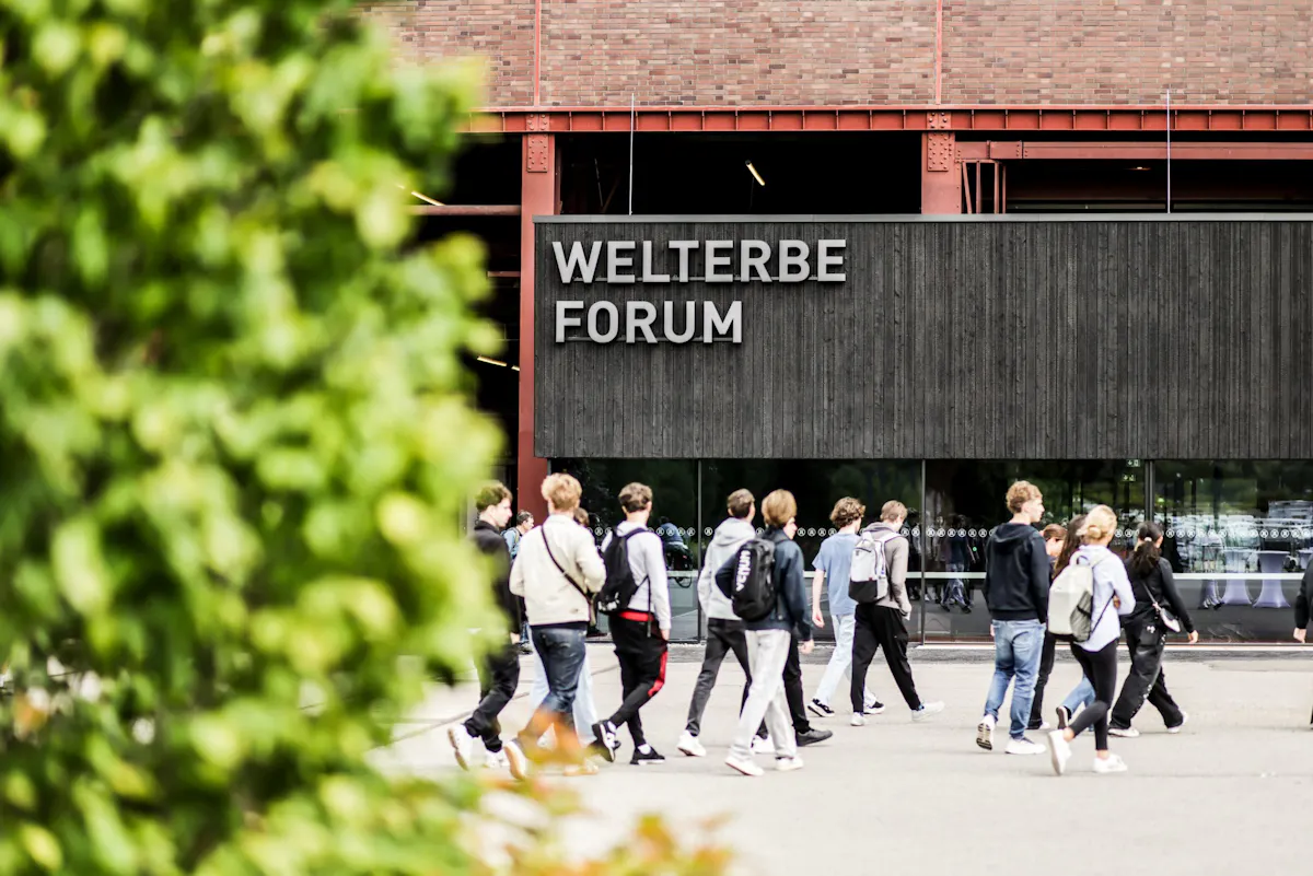 Außenaufnahme des Welterbe Forums auf dem Gelände der Zeche Zollverein. Vor dem schwarzen Eingangsbereich mit großem Schriftzug WELTERBE FORUM geht eine Gruppe junger Menschen über den Platz. Im Vordergrund verdecken unscharfe grüne Blätter einen Teil des Bildes. Dahinter ist die Backsteinfassade mit roten Stahlträgern zu sehen.