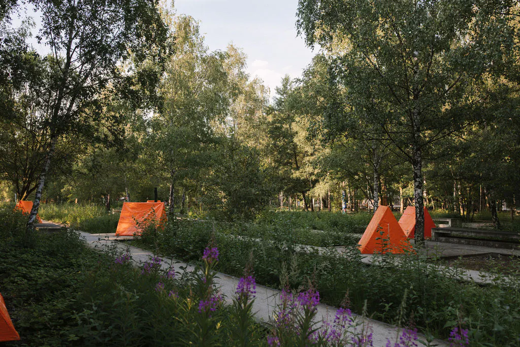 Waldartiger Parkbereich mit Birken, dichtem Grün und schmalen Wegen. Zwischen den Bäumen stehen mehrere orangefarbene, zeltartige Installationen als auffällige Farbakzente. Im Vordergrund blühen violette Pflanzen, das Licht wirkt warm und spätsommerlich.