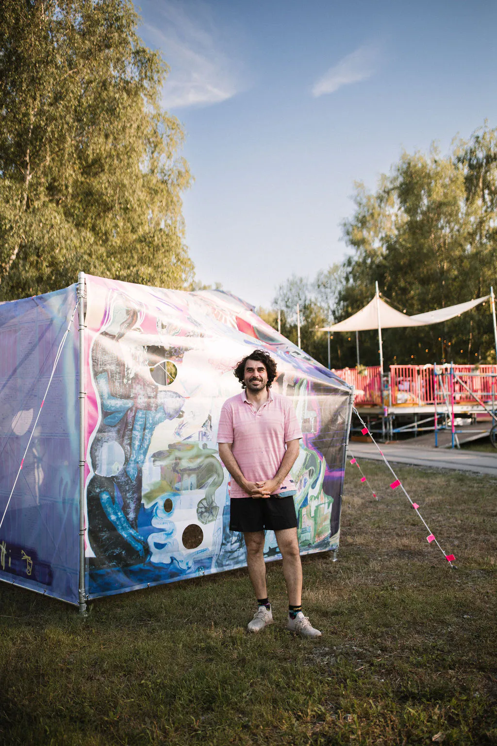 Porträt eines Mannes im Freien vor einer farbig gestalteten, zeltartigen Installation. Die transparente Außenhaut ist mit abstrakten Formen, Farben und runden Öffnungen bemalt. Der Mann trägt ein rosa Poloshirt und kurze schwarze Hose und steht auf einer Wiese. Im Hintergrund sind Bäume, eine helle Überdachung und pinke Absperrungen einer Open-Air-Veranstaltung zu sehen.