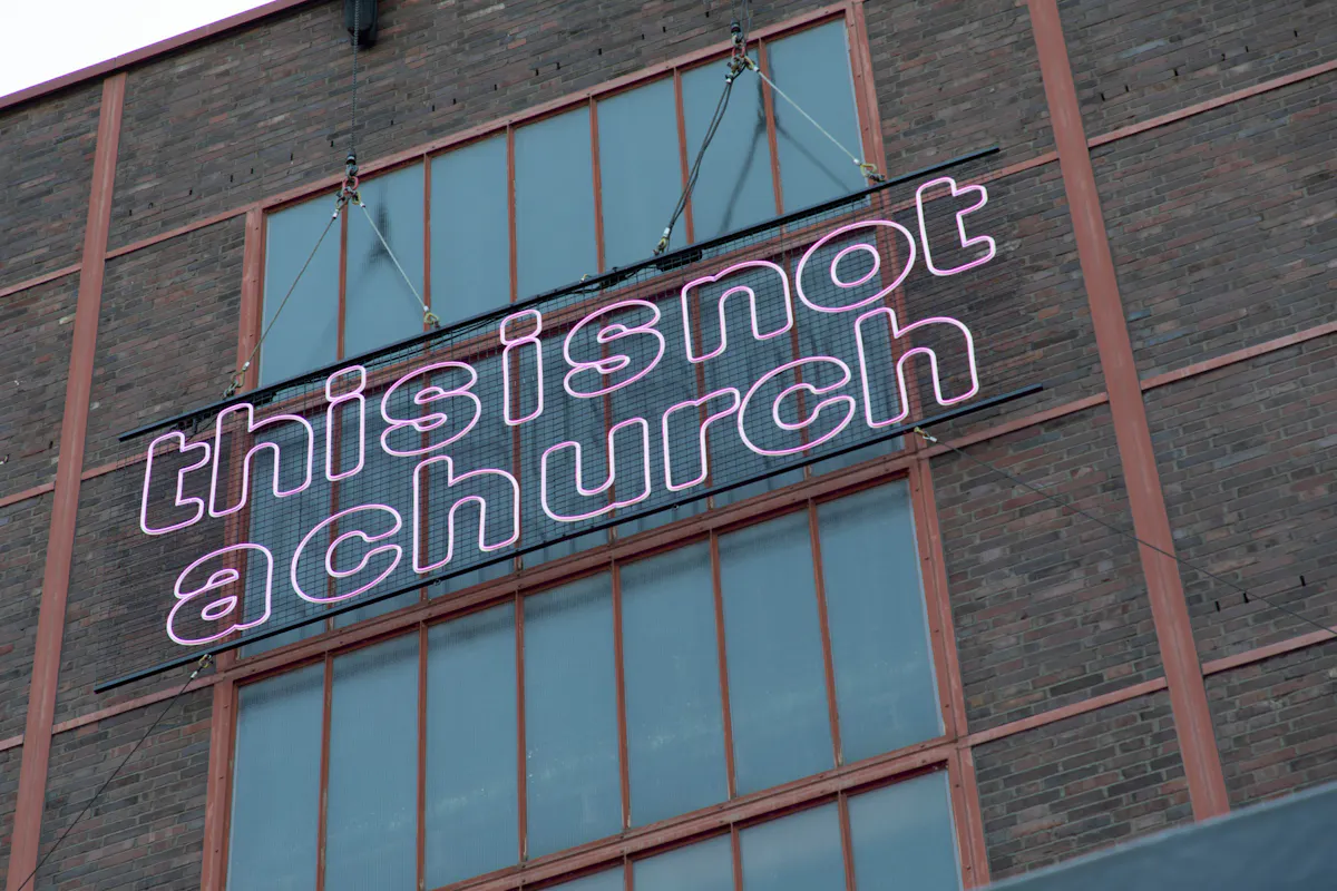 Außenaufnahme einer Backsteinfassade mit Stahlfachwerk und großen Industrie-Fenstern. Davor hängt ein großes Schild oder Leuchtelement mit dem Schriftzug this is not a church in rosa Kontur-Schrift. Das Schild ist an Seilen und Metallstreben befestigt und wirkt wie eine temporäre Kunstinstallation an einem ehemaligen Industriegebäude.