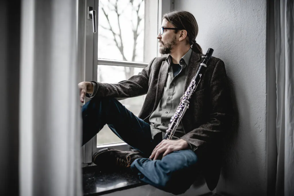 Eine Person mit langen, nach hinten gebundenen Haaren sitzt in lässiger Haltung auf einer Fensterbank. Sie trägt ein Sakko, ein Hemd und eine Jeans. In der rechten Hand hält sie eine Klarinette, die aufrecht an die Schulter gelehnt ist. Im Hintergrund ist ein Fenster mit Blick auf kahle Äste zu sehen. Das Licht fällt weich von draußen herein und erzeugt eine ruhige, nachdenkliche Atmosphäre.