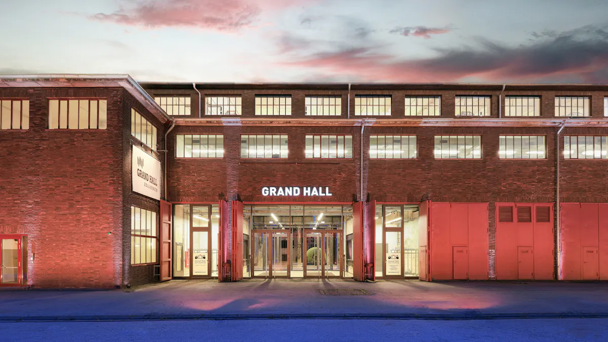 Außenansicht der Grand Hall Zollverein in der Dämmerung. Das große Backsteingebäude hat viele beleuchtete Fenster und einen hell erleuchteten Eingangsbereich mit Glastüren. Über dem Eingang steht der Schriftzug GRAND HALL. Die Fassade wird von warmem rötlichem Licht angestrahlt, während der Boden davor bläulich beleuchtet ist. Im Hintergrund ist ein Abendhimmel mit rosa und grauen Wolken zu sehen.