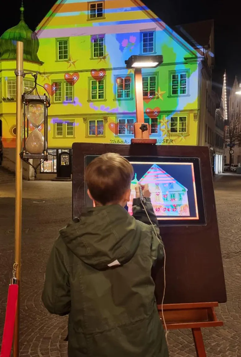 Ein Kind mit hellbraunen Haaren und grüner Jacke steht auf einem nächtlichen Platz und malt mit einem Stift auf einem großen Touchscreen. Auf dem Bildschirm ist die Fassade eines historischen Gebäudes zu sehen. Im Hintergrund wird dieselbe Fassade in leuchtenden Farben und Mustern angestrahlt. Links im Bild steht eine große Sanduhr auf einem Holzständer. Die Szene ist von Straßenlaternen beleuchtet, und der Platz ist menschenleer.