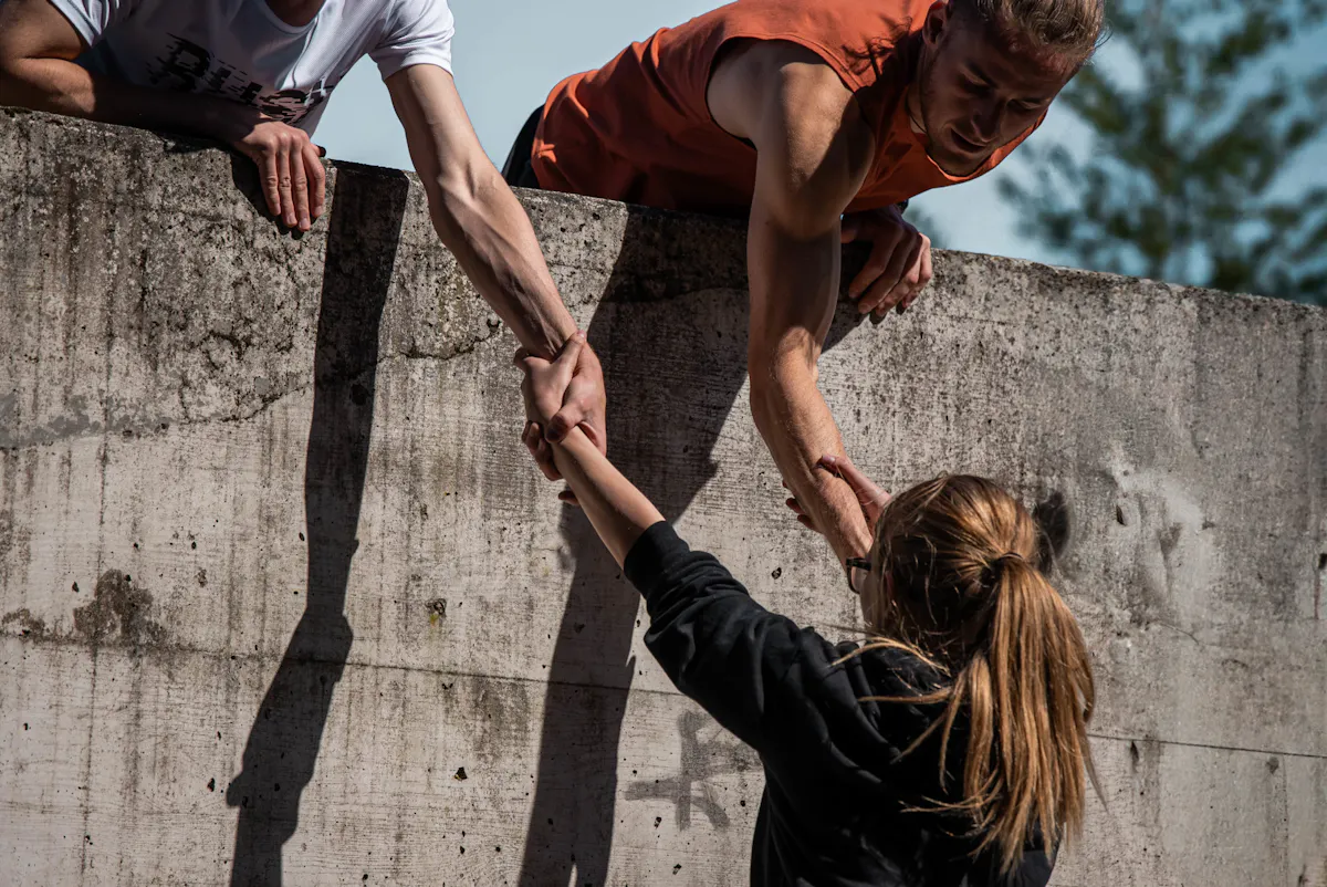 Eine Person mit Sonnenbrille und dunkler Jacke steht vor einer Betonmauer und greift nach der Hand einer anderen Person, die sich über die Mauer lehnt. Eine weitere Person im weißen T-Shirt steht daneben und hält ebenfalls die Hand der Person auf der Mauer. Die Szene spielt im Freien bei Tageslicht.