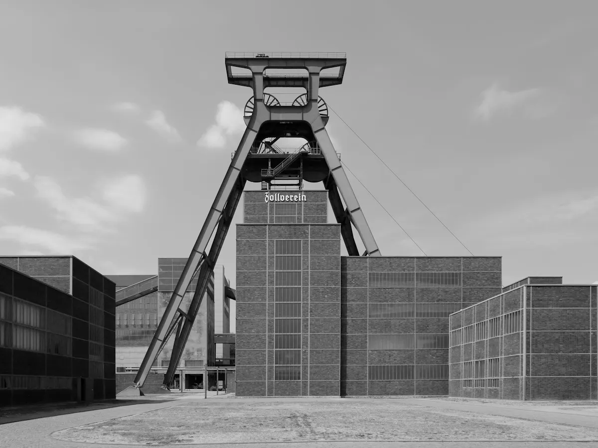 Schwarz-Weiß-Aufnahme des Doppelbock-Fördergerüsts der Zeche Zollverein in frontaler Perspektive. Die markante Stahlkonstruktion erhebt sich hinter mehreren klar gegliederten Backsteingebäuden. Auf dem mittleren Gebäude steht der Schriftzug Zollverein. Im Vordergrund liegt eine freie Platzfläche, darüber ein heller Himmel mit wenigen Wolken.