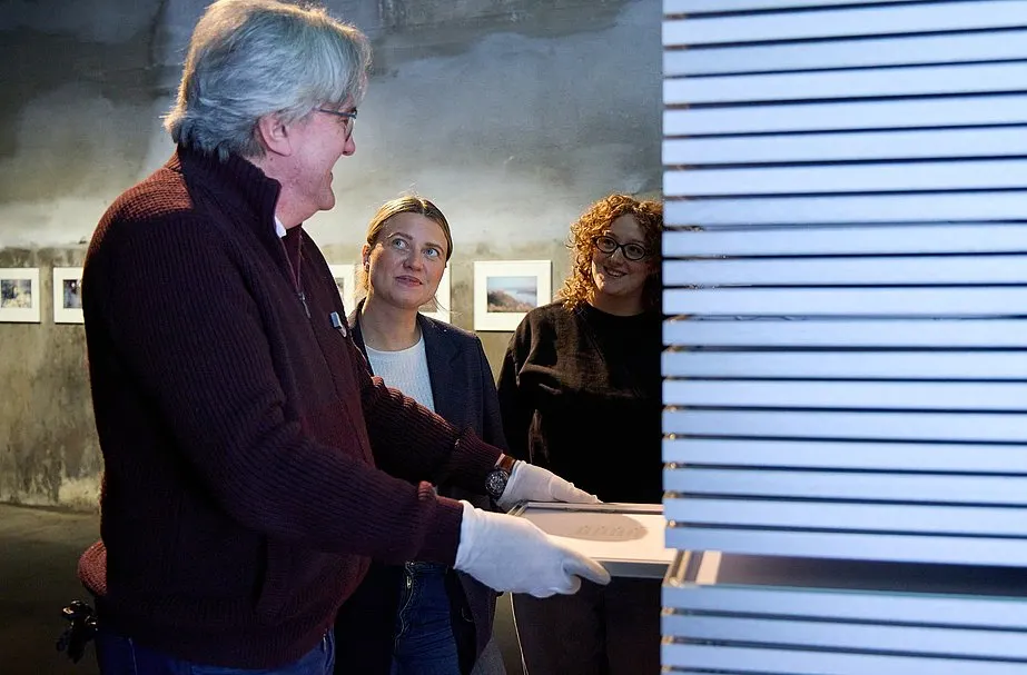 Ausstellungsszene mit drei Personen vor einer großen weißen Lamellenstruktur. Ein älterer Mann mit grauen Haaren, Brille und weißen Handschuhen hält oder führt ein flaches Objekt in die Installation ein. Zwei Frauen stehen daneben und schauen aufmerksam zu. Im Hintergrund sind raue Wände und gerahmte Bilder zu sehen.