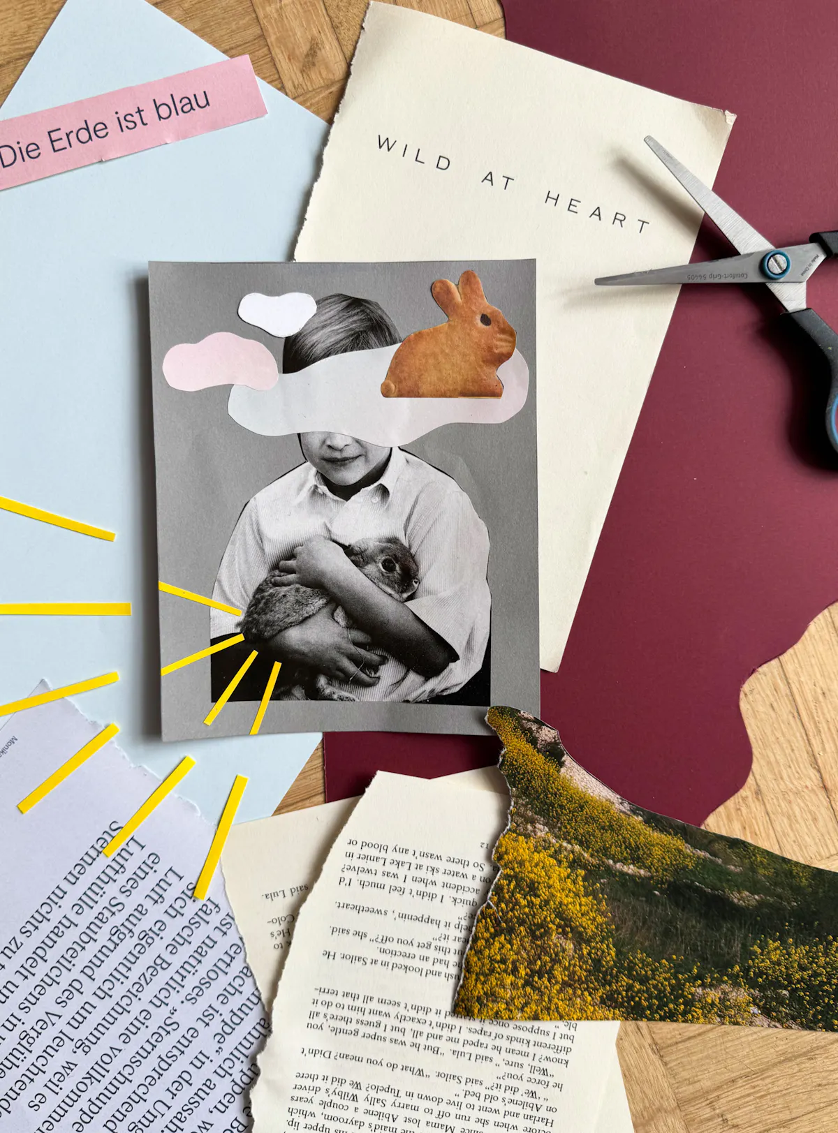 Collage aus Papier, Fotos und Bastelmaterialien auf einem Holztisch. In der Mitte liegt ein schwarz-weißes Bild eines Kindes mit einem Kaninchen im Arm, ergänzt durch ausgeschnittene Wolkenformen, ein braunes Hasenmotiv und gelbe Papierstreifen wie Sonnenstrahlen. Darum herum liegen farbige Papiere, Textausschnitte, ein Landschaftsausschnitt, eine Schere und Blätter mit den Aufschriften Die Erde ist blau und Wild at Heart.