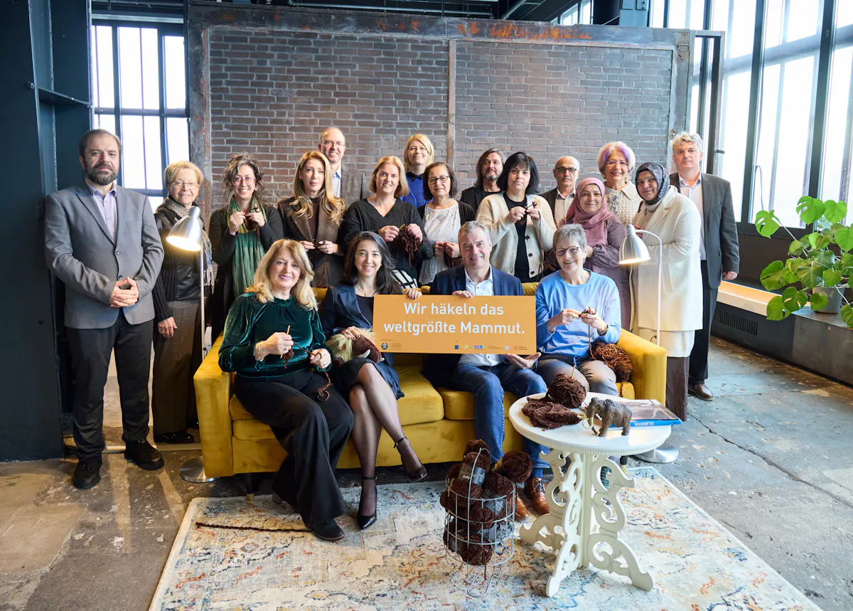 Gruppenfoto in einem industriellen Innenraum mit Backsteinwand, großen Fenstern und gelbem Sofa. Mehrere Personen stehen und sitzen zusammen, einige halten Häkelnadeln oder braunes Garn in den Händen. In der Mitte wird ein Schild mit der Aufschrift Wir häkeln das weltgrößte Mammut. gehalten. Auf einem kleinen Tisch liegen weitere Garnknäuel und eine kleine Mammutfigur.