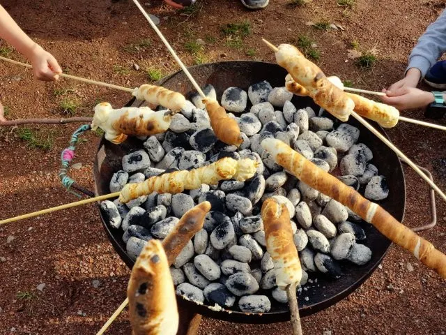 Mehrere Hände halten lange Holzstöcke mit aufgerolltem Brotteig über eine runde Feuerschale mit glühender Holzkohle. Der Teig ist an einigen Stellen goldbraun gebacken, an anderen noch hell. Die Szene spielt sich im Freien auf erdigem Boden ab.